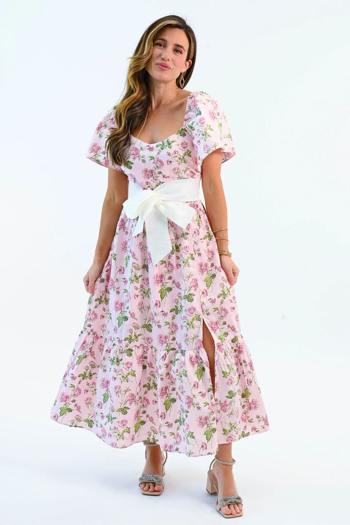 A woman wearing a pink floral puff sleeve maxi dress with a waist tie belt and side slit, styled with elegant heels