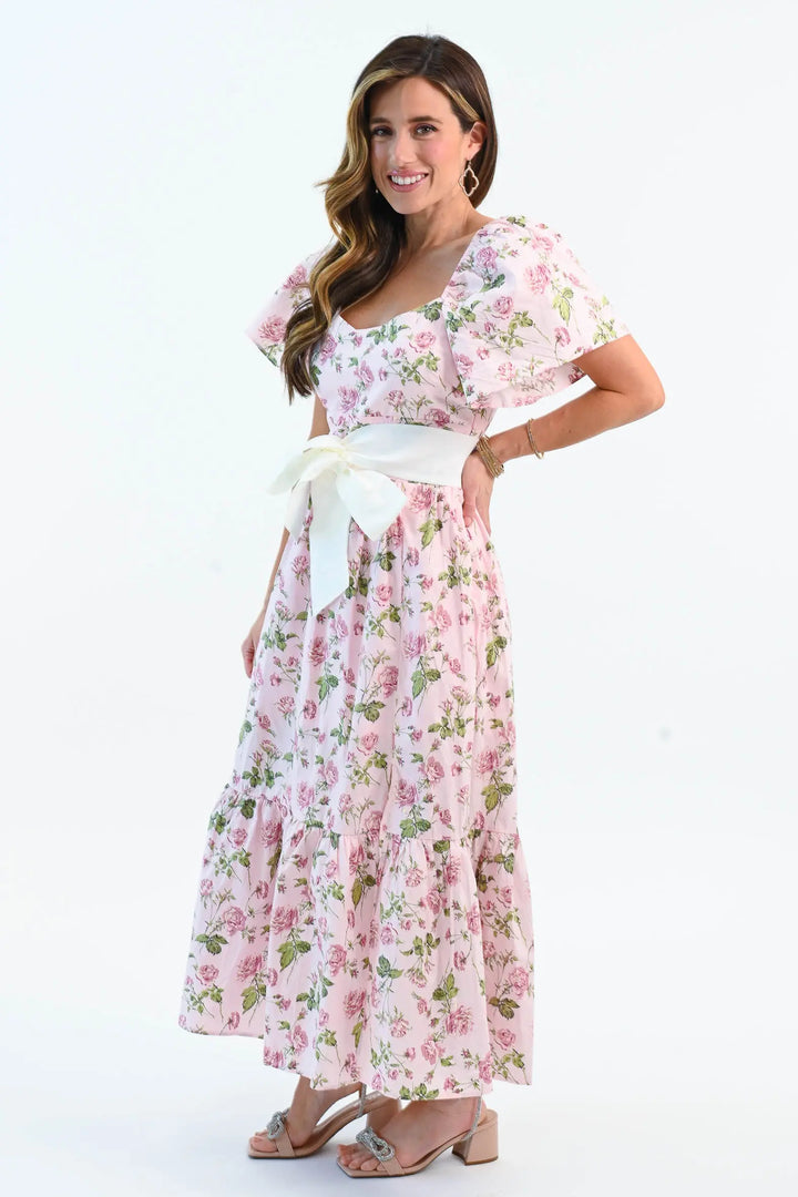 A woman wearing a pink floral puff sleeve maxi dress with a waist tie belt and side slit, styled with elegant heels