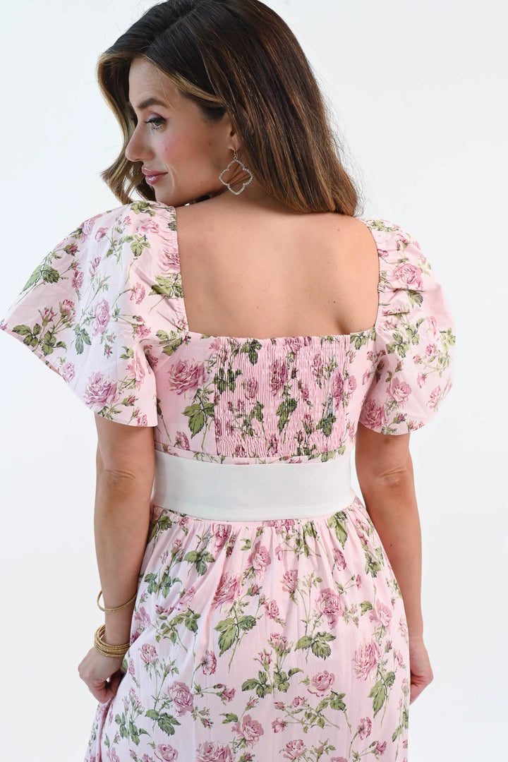A woman wearing a pink floral puff sleeve maxi dress with a waist tie belt and side slit, styled with elegant heels