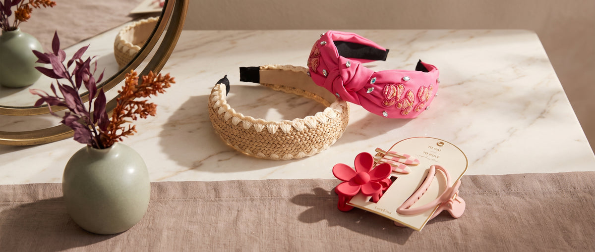 A braided beige headband, a pink embellished knotted headband and a set of hair clips on a marble dressing table