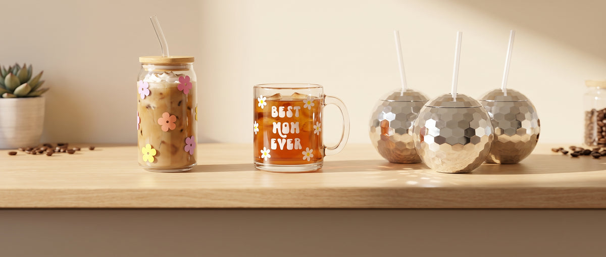 A daisy print glass cup with bamboo lid, a Best Mom Ever glass mug and silver disco ball cups on a sunny oak café counter