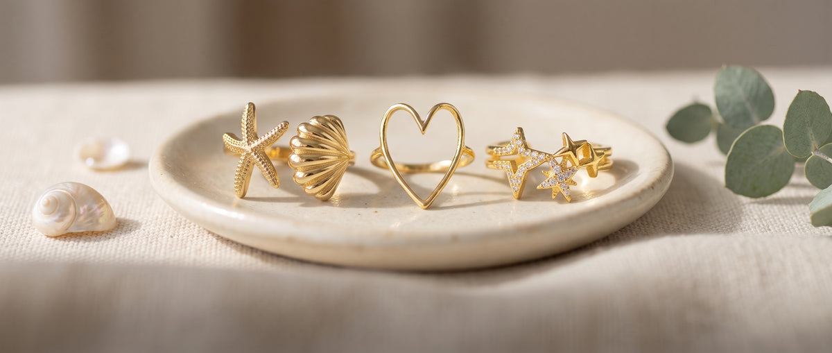 A gold starfish and seashell ring, a gold heart ring and a gold star ring on a ceramic dish on cream linen with shells and eucalyptus
