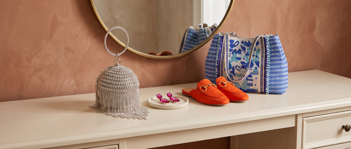 A rhinestone fringe ball bag, pink crystal drop earrings, orange suede mules and a blue embroidered tote on a cream dressing table under a round mirror