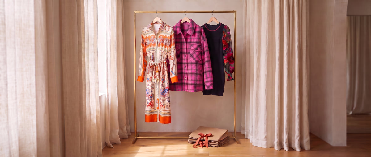 Three sale pieces on a brass rack between linen curtains: an orange-bordered scarf-print tie-waist shirt dress, a pink and black plaid shacket and a black sweater dress with sheer floral sleeves