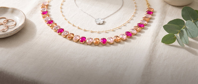 A silver flower pendant necklace, a fine gold pearl chain and a pink and peach gem statement necklace laid on cream linen with eucalyptus
