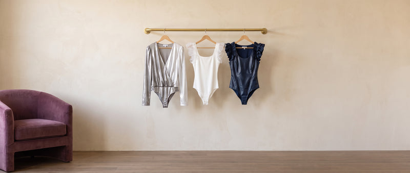 A silver metallic wrap bodysuit, a white ruffle-shoulder bodysuit and a navy faux leather ruffle bodysuit on a brass rail beside a plum chair