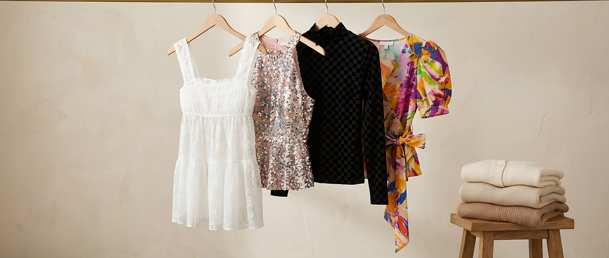 A white lace smocked top, a sequin halter top, a black velvet checker top and a bright printed wrap top on wooden hangers beside a stool of folded knits