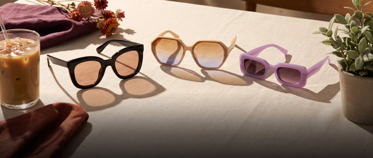Black, cream and lilac oversized sunglasses on a sunlit café table beside an iced coffee