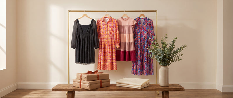 Black glitter, orange print, pink tiered suede and purple print dresses on a brass rail above a wooden bench with wrapped gifts