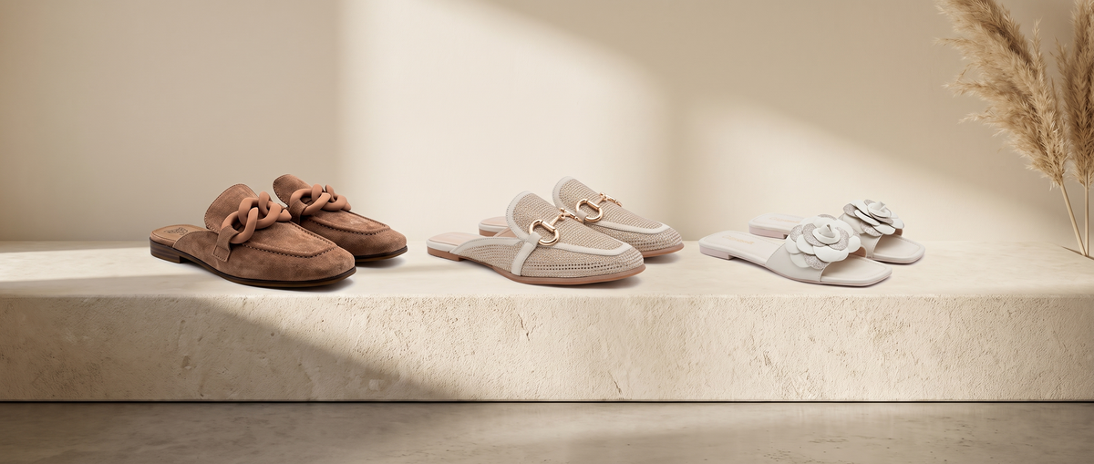 Brown suede chain mules, beige perforated buckle mules and white flower slide sandals on a cream stone plinth with dried pampas