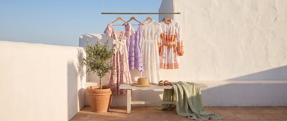A pink floral bow-belt maxi dress, a lilac crochet mini, a white lace midi and a floral border mini dress on a brass rail on a sunny white terrace, with a straw hat and sandals on a bench