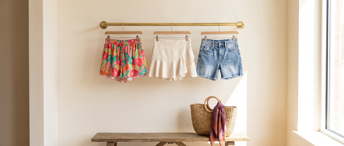 Floral shorts, a cream ruffle skort and denim shorts hanging on a brass rail above a bench with a straw bag
