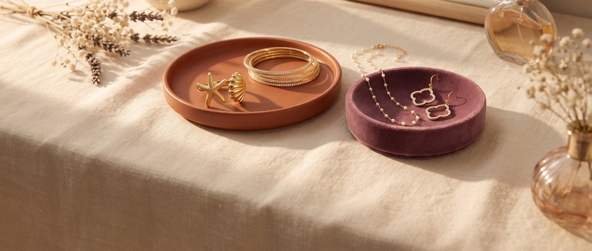 Gold starfish ring and bangles in a terracotta dish beside a pearl chain necklace and clover earrings in a plum velvet dish on linen