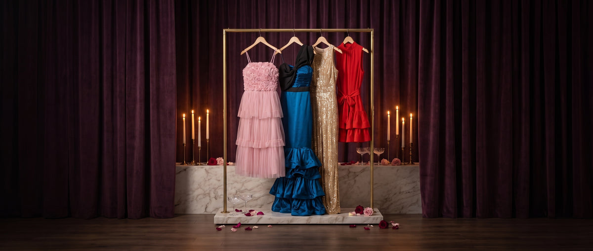 Pink tulle, blue satin, gold sequin and red ruffle party dresses on a gold rail in front of plum velvet curtains with candles