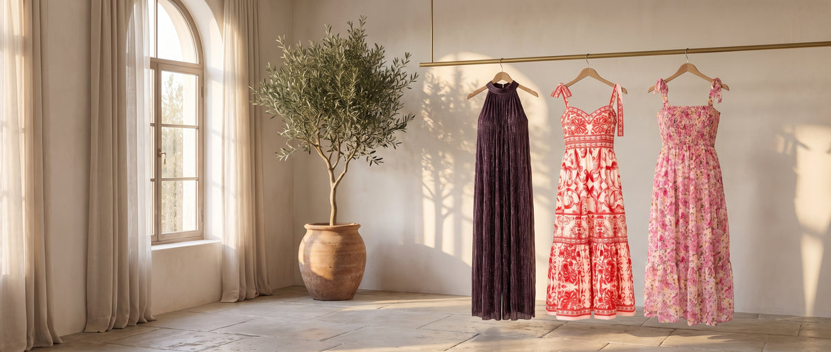 Plum pleated halter jumpsuit, red tile-print maxi and pink floral smocked maxi on a brass rail in a sunlit room with an olive tree