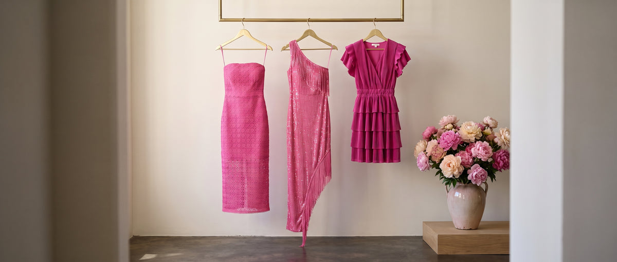 Three hot pink dresses, lace, sequin fringe and tiered, hanging on a brass rail beside a vase of peonies