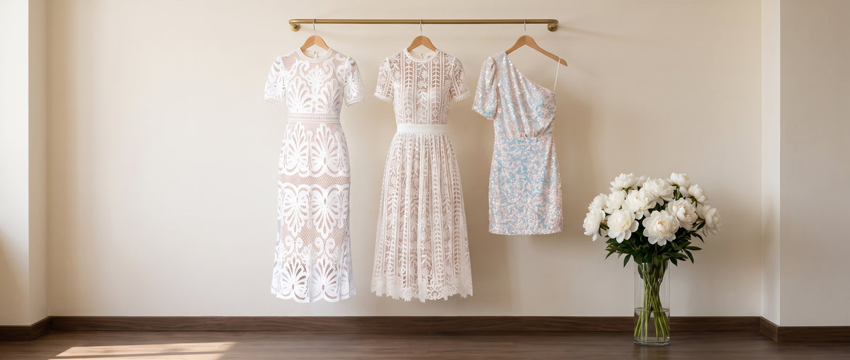 Two white lace midi dresses and an iridescent sequin one-shoulder mini dress on a brass rail beside a vase of white peonies
