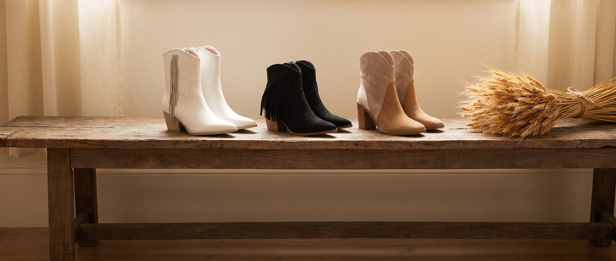 White rhinestone western boots, black fringe ankle boots and two-tone suede booties on a rustic wooden bench with dried wheat