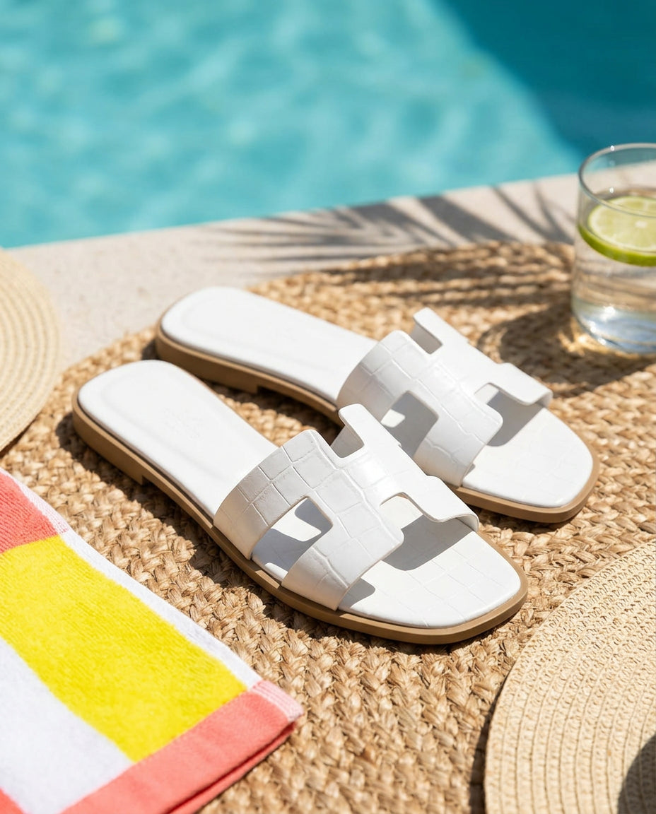 White flat slide sandals styled poolside in a bright playful summer scene, framed with woven textures and vacation color accents, no text in frame.  (sources: 1)