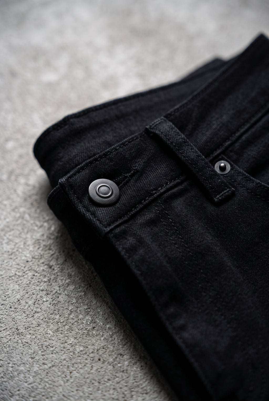 Close detail of the black jeans waistband, button, belt loop, pocket rivet and tonal stitching.