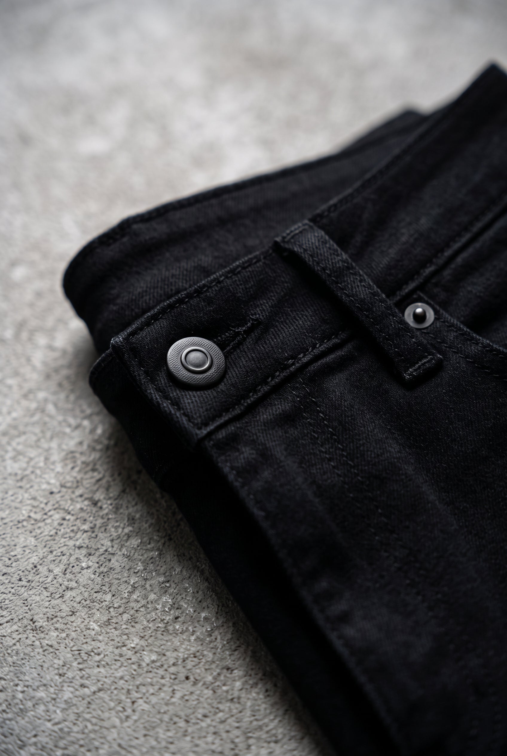 Close detail of the black jeans waistband, button, belt loop, pocket rivet and tonal stitching.