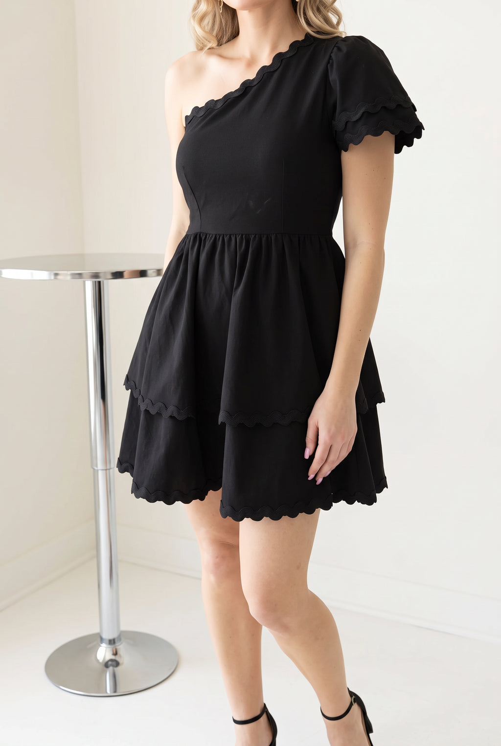Black one-shoulder tiered mini dress styled with minimal heels for a cocktail party look.