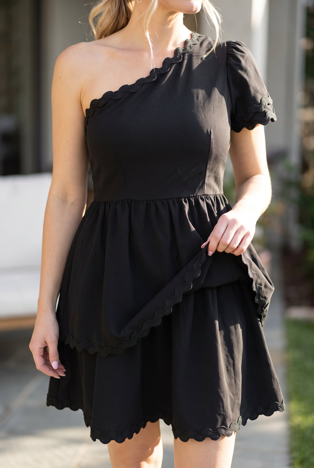 Close view of the black polyester dress showing its gathered tiers, wavy braid trim, and soft drape.
