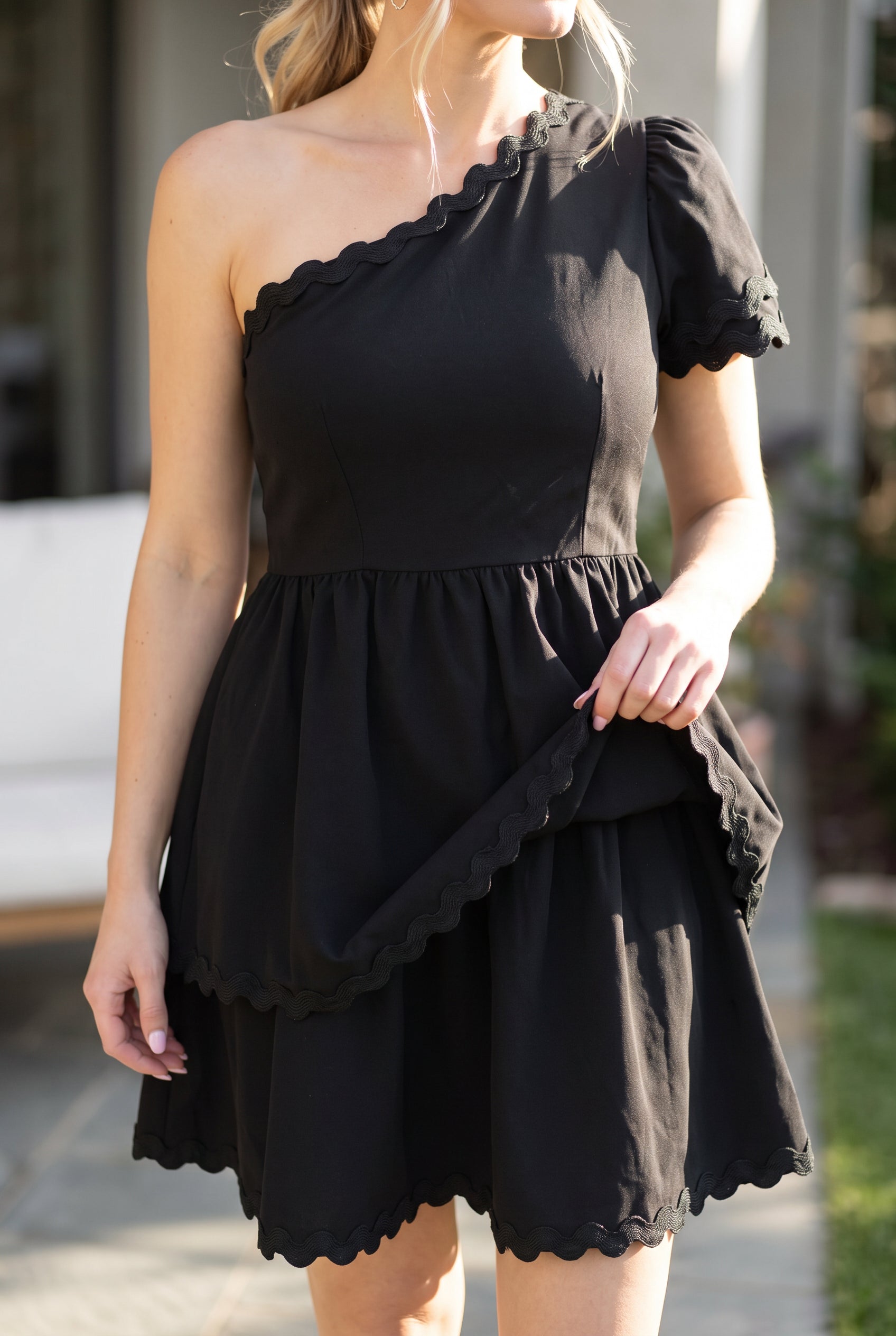Close view of the black polyester dress showing its gathered tiers, wavy braid trim, and soft drape.