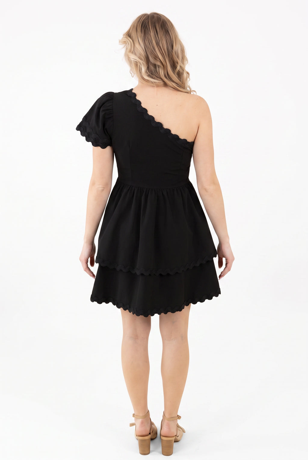 Back view of the black mini dress showing its low asymmetric neckline, fitted waist, and tiered skirt.
