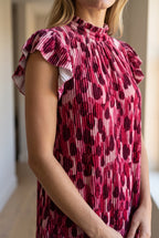 Close view of the pink dress showing vertical pleats, velvet-like sheen and layered flutter sleeve.