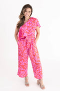Model wearing a vibrant pink and coral floral one-shoulder ruffle jumpsuit with wide-leg pants, styled with open-toe heels.