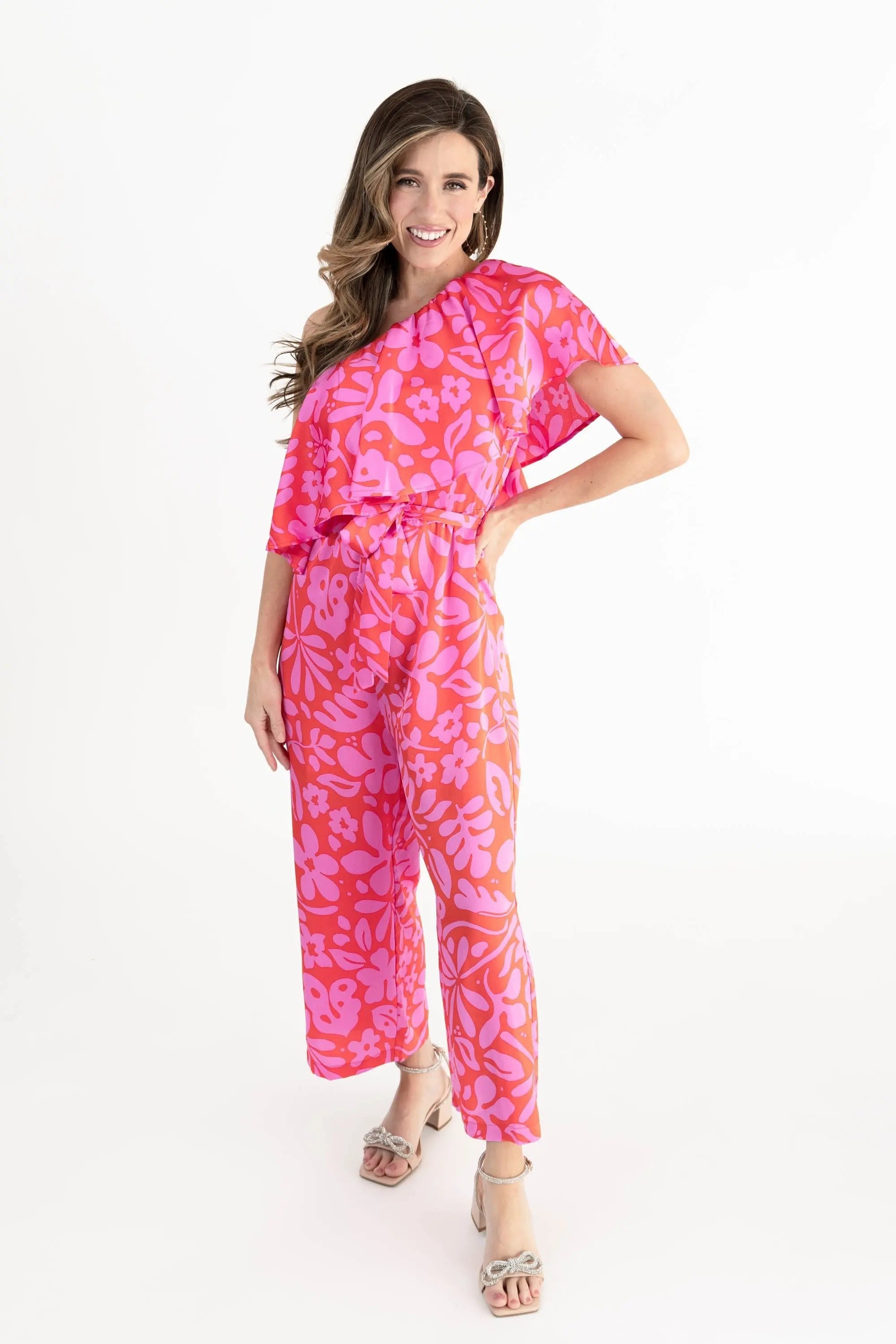 Model wearing a vibrant pink and coral floral one-shoulder ruffle jumpsuit with wide-leg pants, styled with open-toe heels.