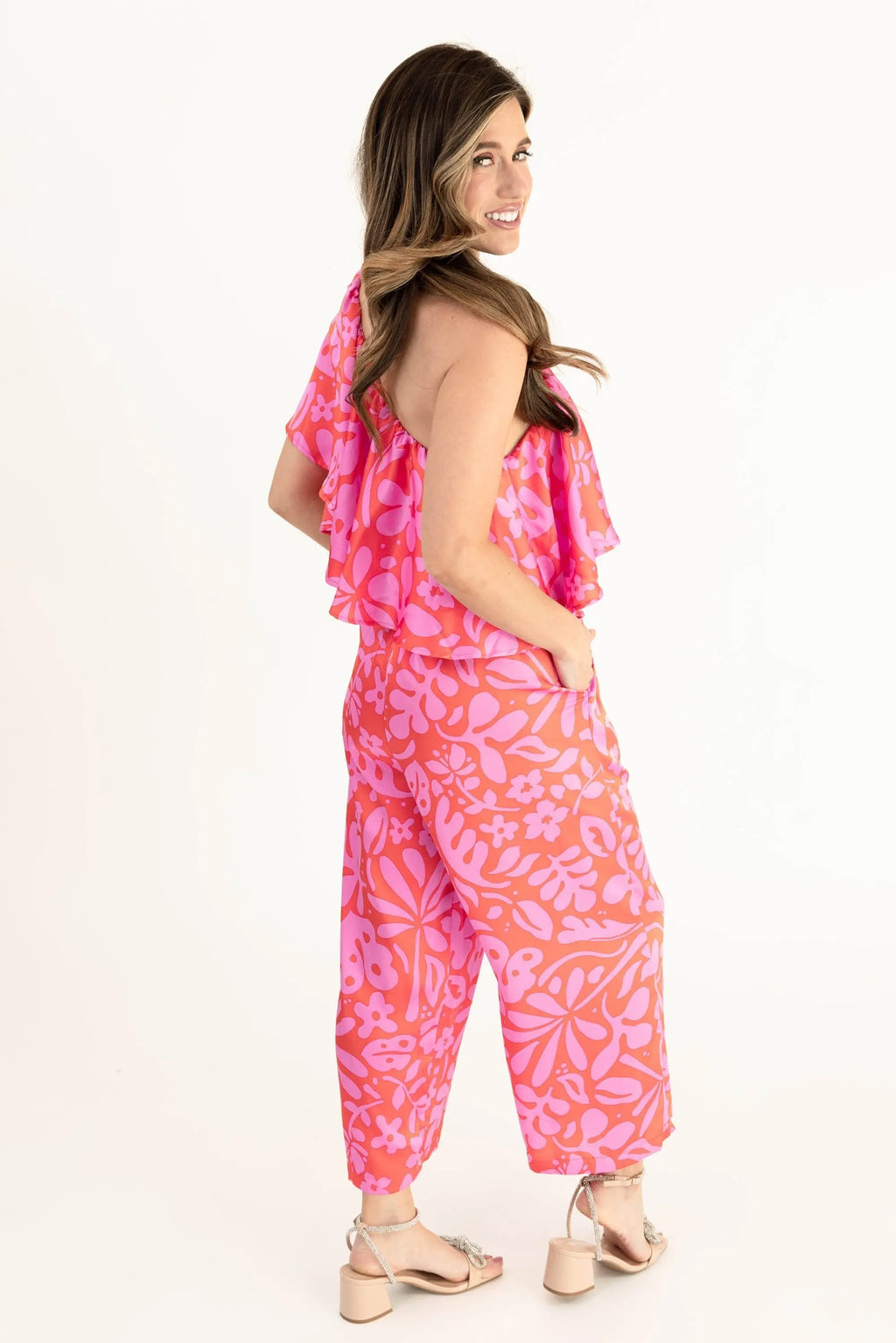 Model wearing a vibrant pink and coral floral one-shoulder ruffle jumpsuit with wide-leg pants, styled with open-toe heels.
