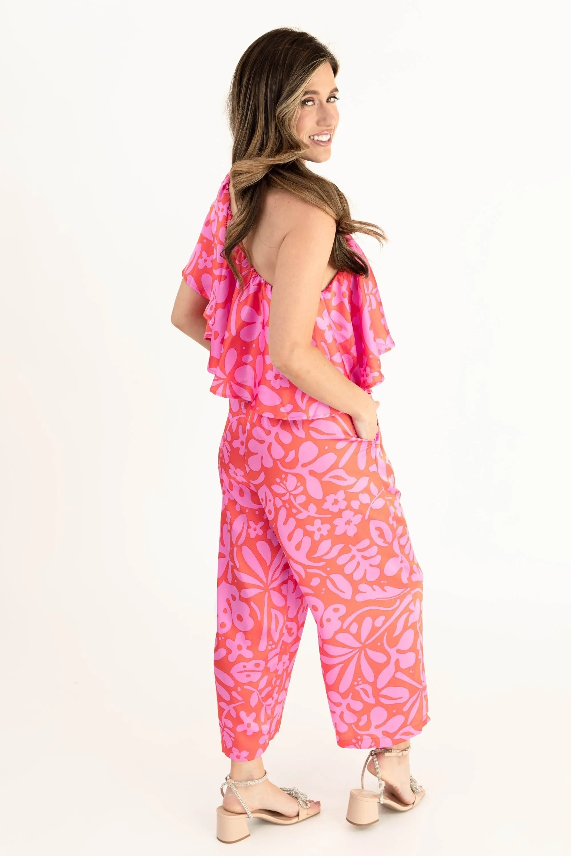 Model wearing a vibrant pink and coral floral one-shoulder ruffle jumpsuit with wide-leg pants, styled with open-toe heels.