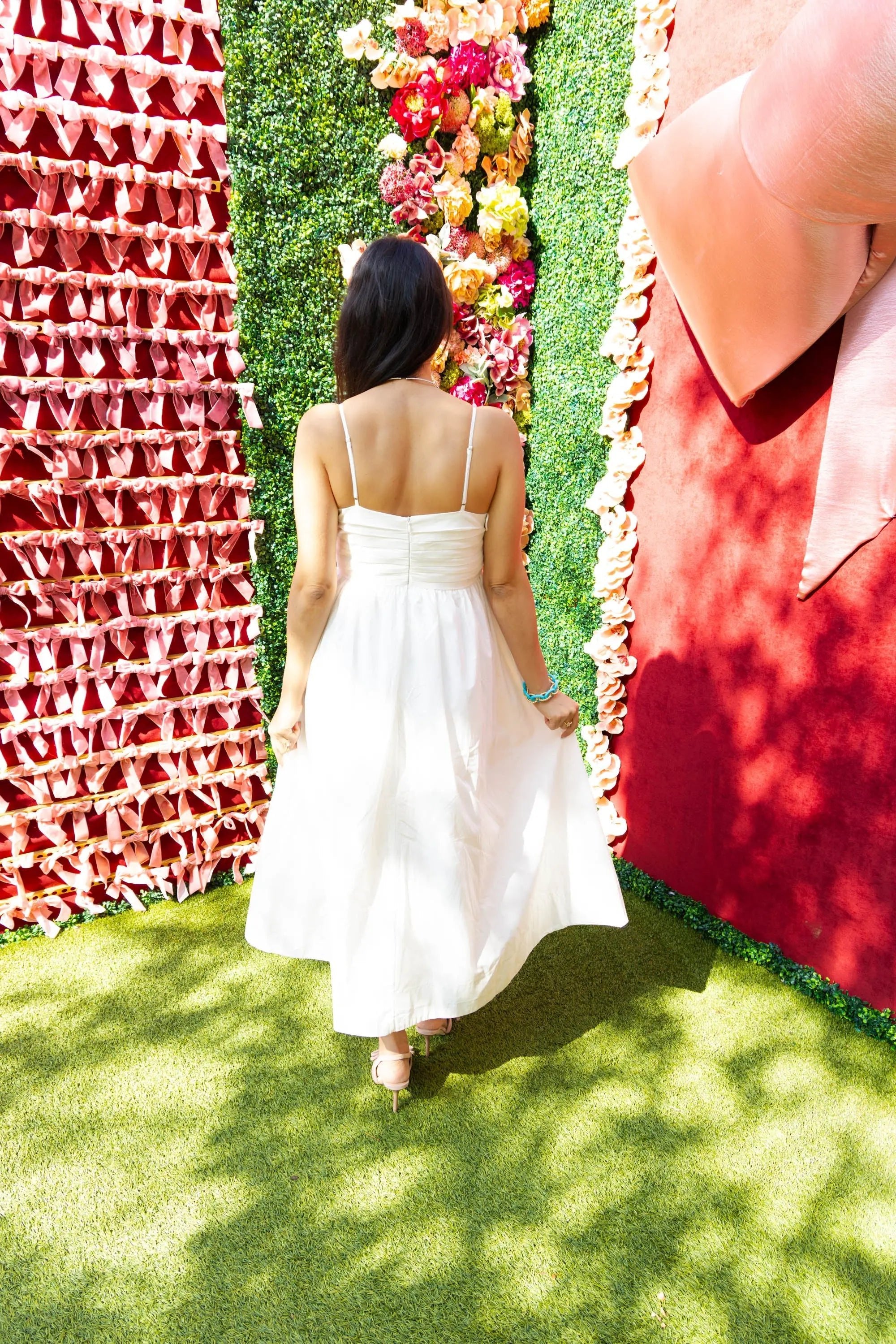 A woman wearing a white pleated midi dress with spaghetti straps, holding a quilted handbag, styled with nude heels, standing in front of a pink ribbon wall.