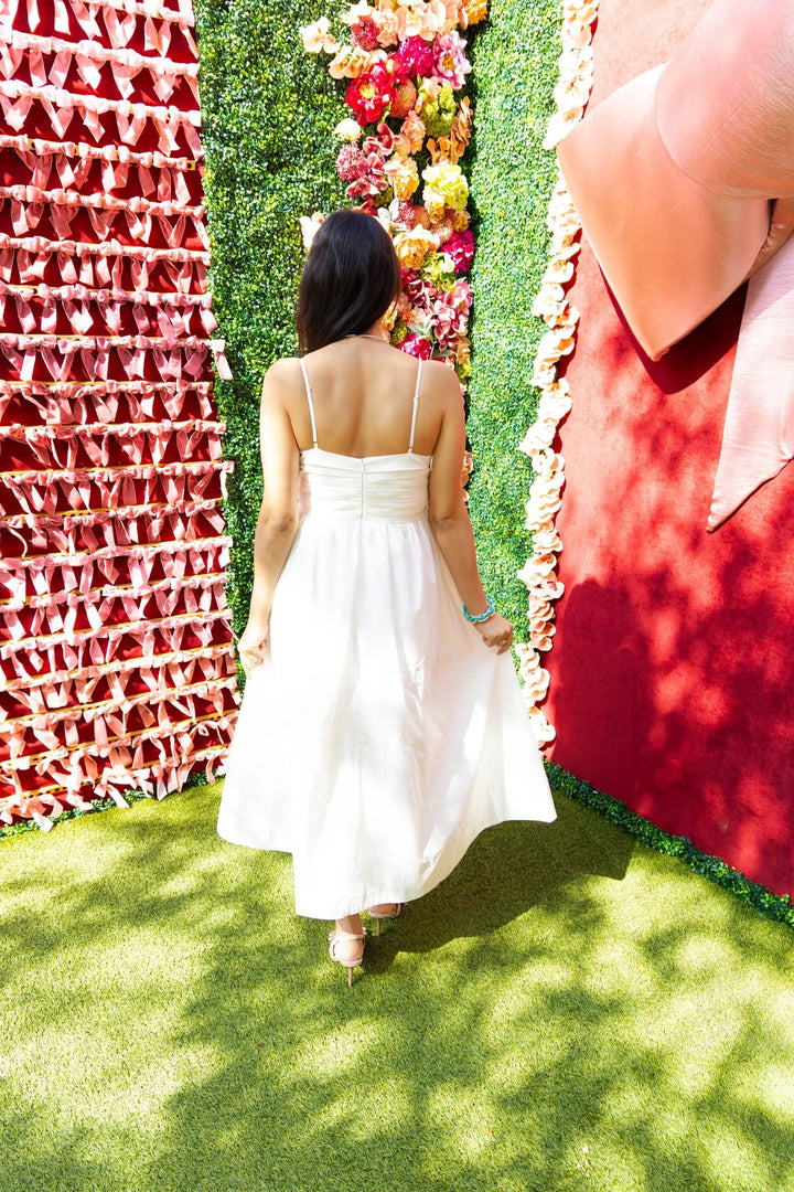 A woman wearing a white pleated midi dress with spaghetti straps, holding a quilted handbag, styled with nude heels, standing in front of a pink ribbon wall.