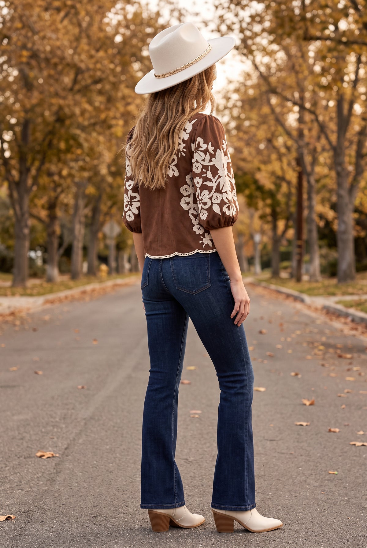 Ivory felt fedora with a braided gold chain band worn with a brown embroidered faux suede top, dark flare jeans and cream western booties on an autumn street, seen from behind