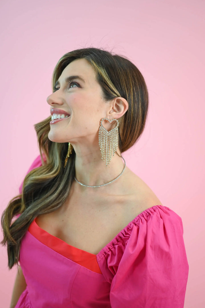 Woman wearing radiant heart fringe rhinestone earrings with cascading rhinestones, styled with an off-shoulder pink dress.