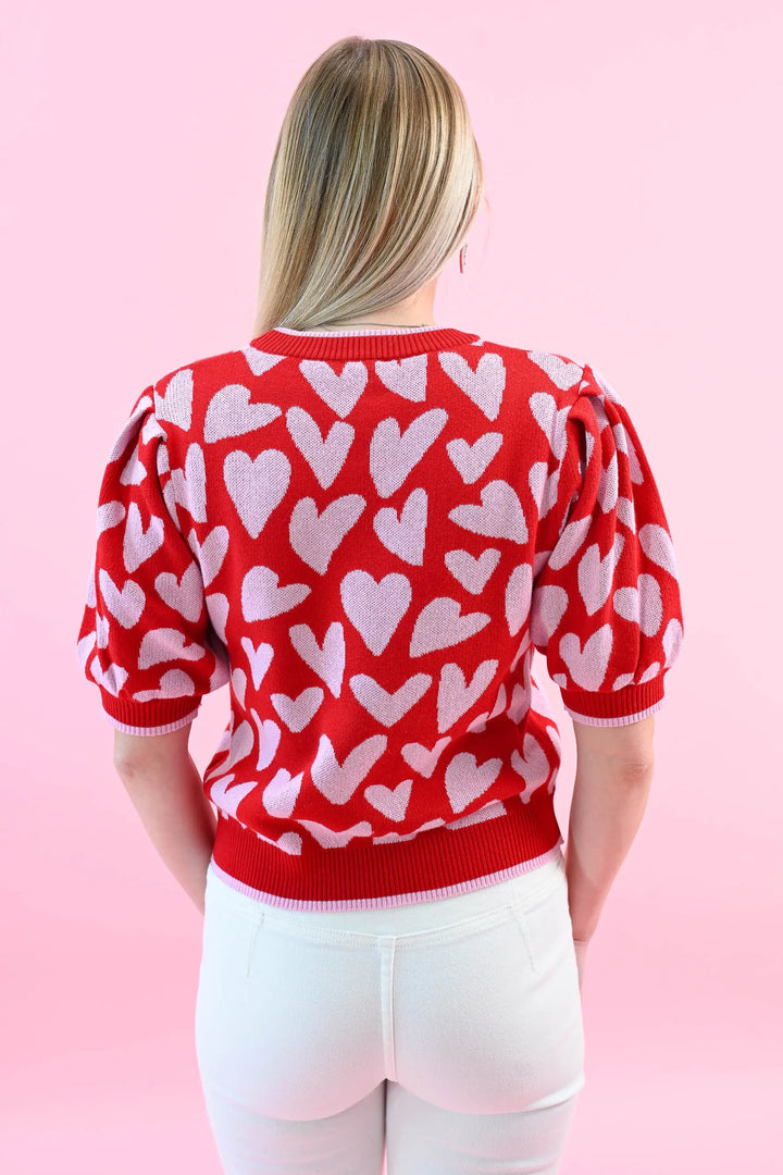 Woman wearing a red heart print puff sleeve sweater with white jeans, perfect for a chic and cozy Valentine's Day outfit.