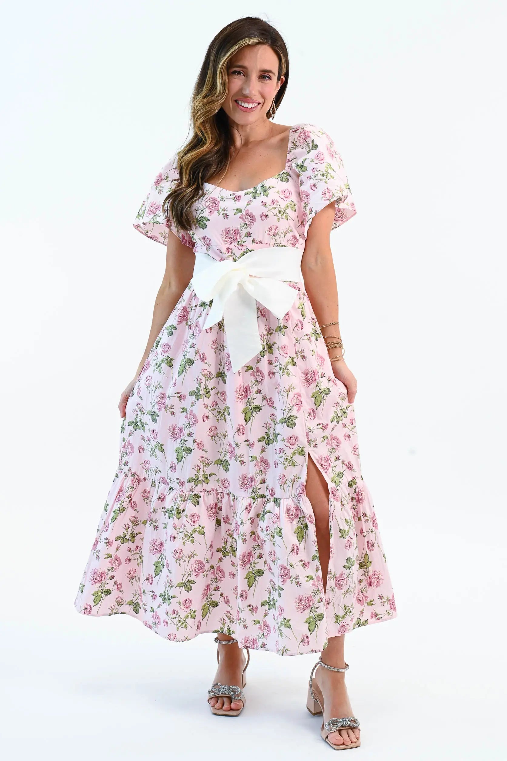 A woman wearing a pink floral puff sleeve maxi dress with a waist tie belt and side slit, styled with elegant heels