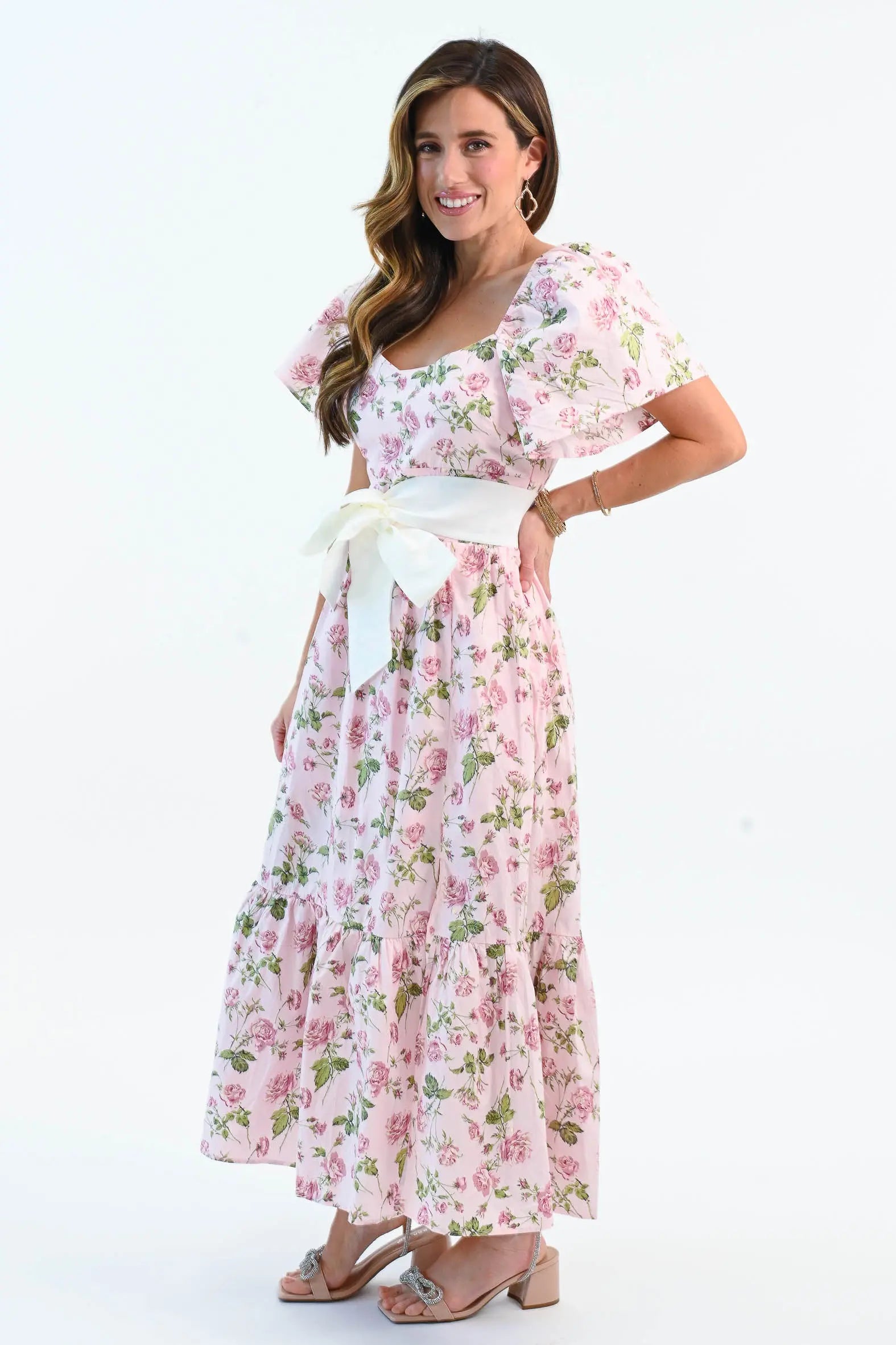 A woman wearing a pink floral puff sleeve maxi dress with a waist tie belt and side slit, styled with elegant heels