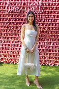 A woman wearing a white pleated midi dress with spaghetti straps, holding a quilted handbag, styled with nude heels, standing in front of a pink ribbon wall.