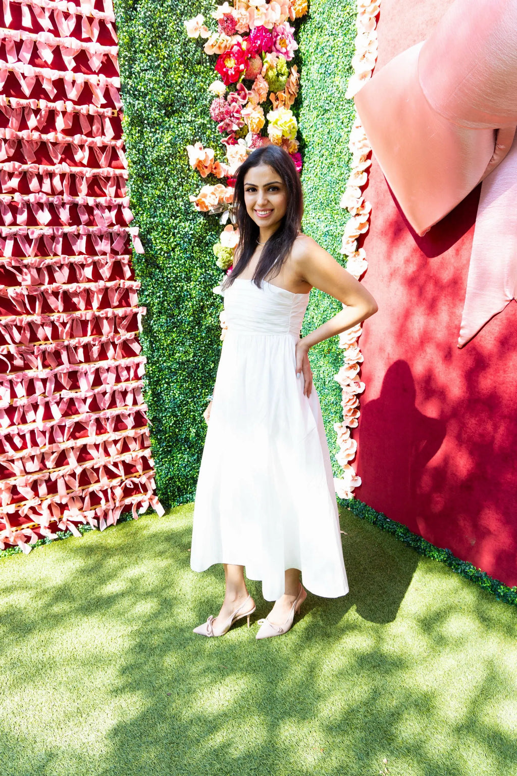A woman wearing a white pleated midi dress with spaghetti straps, holding a quilted handbag, styled with nude heels, standing in front of a pink ribbon wall.