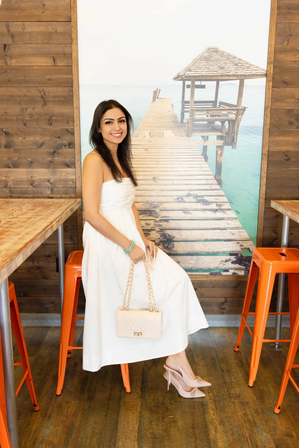 A woman wearing a white pleated midi dress with spaghetti straps, holding a quilted handbag, styled with nude heels, standing in front of a pink ribbon wall.