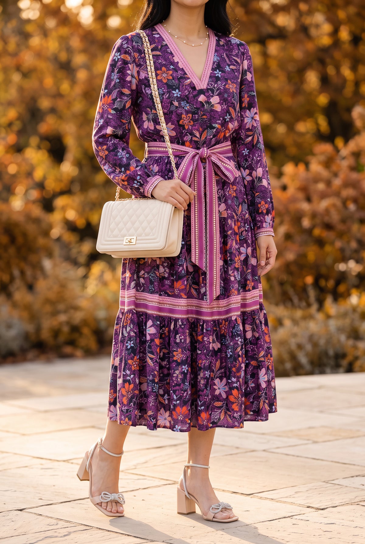Purple floral long sleeve midi dress with a pink sash, ivory quilted chain shoulder bag, gold pearl chain necklace and nude rhinestone bow block heel sandals in an autumn garden
