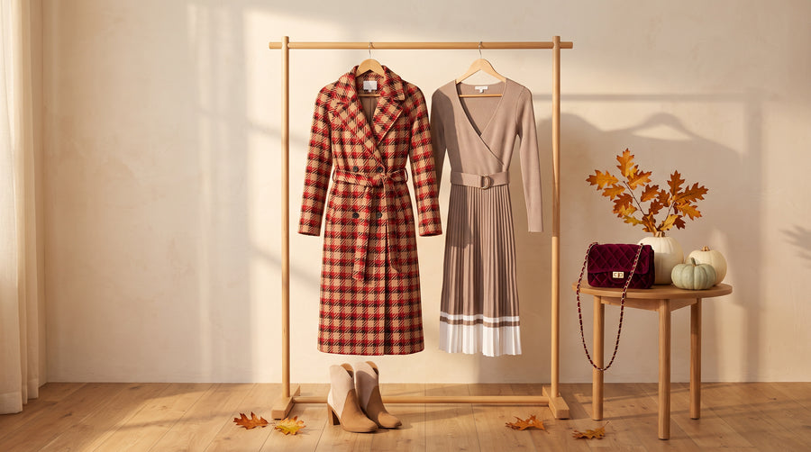 A red plaid belted coat and a taupe pleated sweater dress hanging on an oak rail in golden autumn light, with suede ankle booties below and a burgundy velvet crossbody bag beside white pumpkins and oak leaves