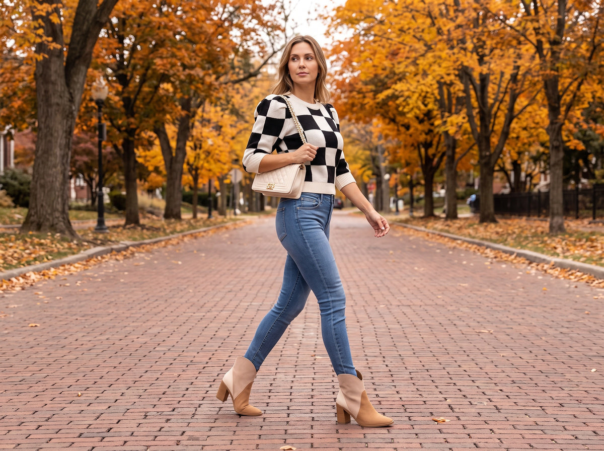 A woman in a black and white checkered puff-sleeve sweater, blue skinny jeans, tri-tone booties and a cream quilted bag, on a brick autumn street