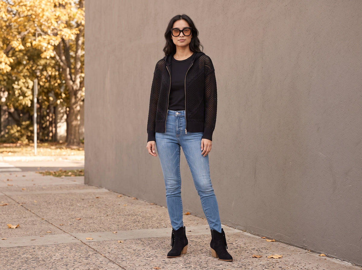 A woman in a black crochet zip hoodie, skinny jeans, black fringe ankle boots and dark sunglasses by a grey wall