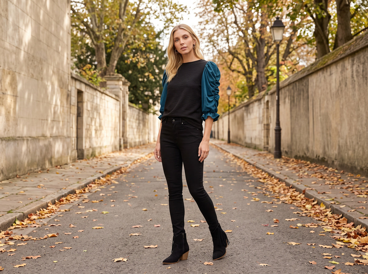 A woman in a black knit top with teal puff sleeves, black jeans and black fringe ankle boots