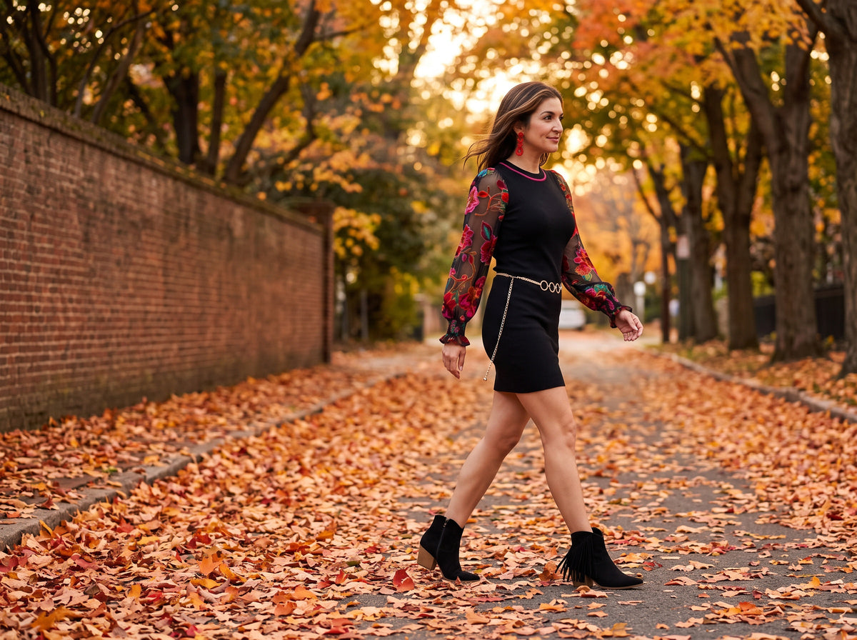 A woman in a black mini sweater dress with sheer floral sleeves, a gold chain belt, red earrings and black fringe ankle boots, on a leafy autumn lane